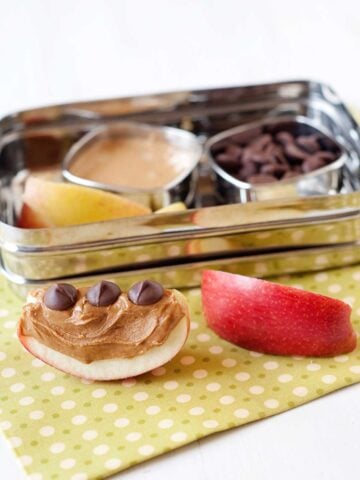 two apple slices with peanut butter and chocolate chips. Behind, a lunchbox with more apples, pb, and chocolate chips