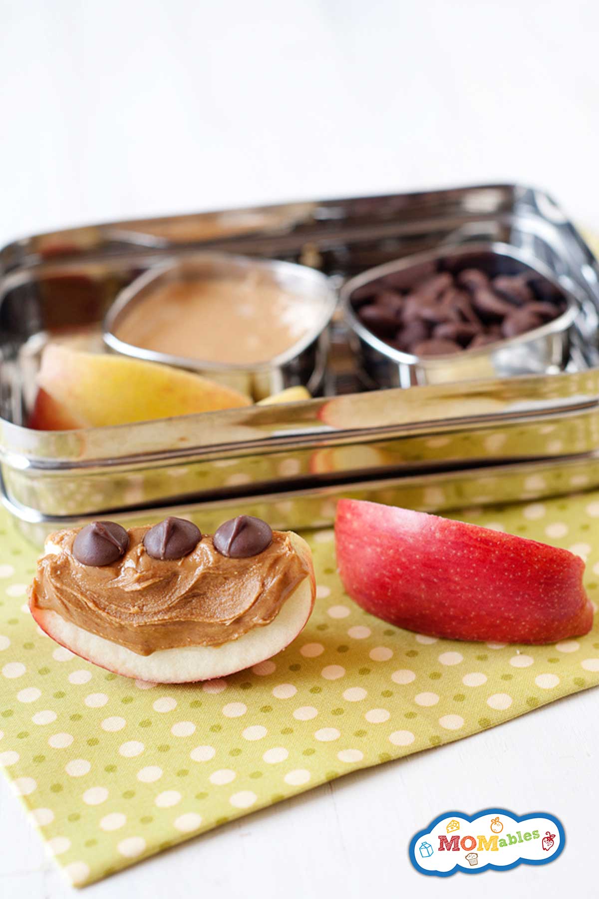 two apple slices with peanut butter and chocolate chips. Behind, a lunchbox with more apples, pb, and chocolate chips