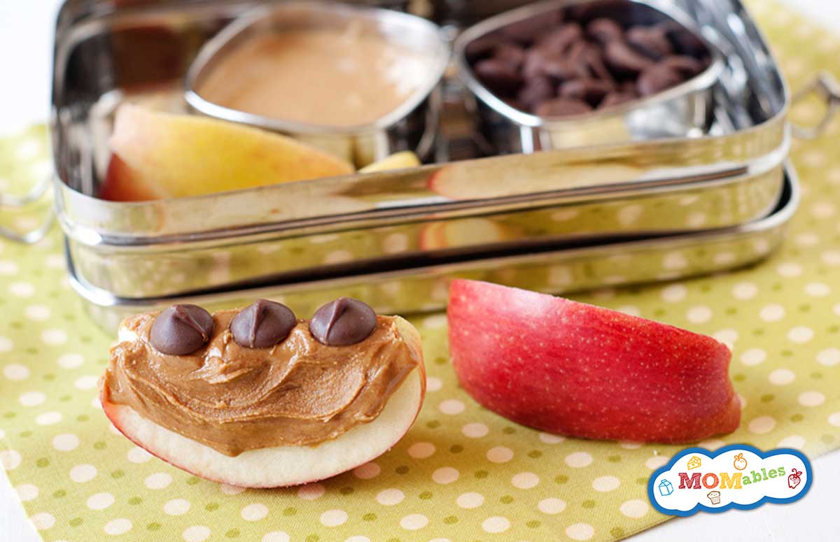 two apple slices with peanut butter and chocolate chips. Behind, a lunchbox with more apples, pb, and chocolate chips
