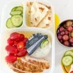 greek chicken bento box with cherry tomatoes, sliced cucumber, pita and tzatziki sauce, plus grapes on a small container on the side.