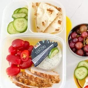 greek chicken bento box with cherry tomatoes, sliced cucumber, pita and tzatziki sauce, plus grapes on a small container on the side.