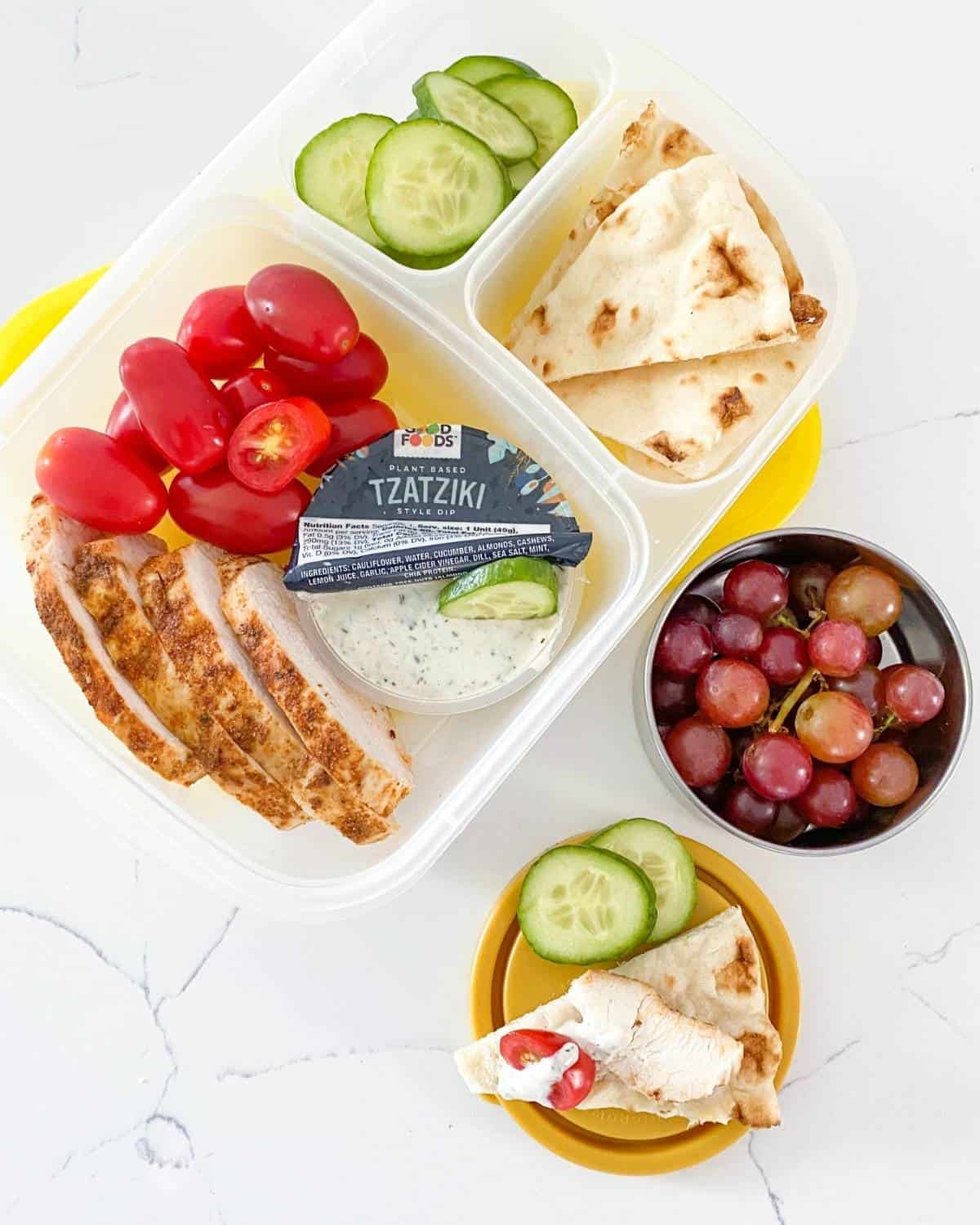 greek chicken bento box with cherry tomatoes, sliced cucumber, pita and tzatziki sauce, plus grapes on a small container on the side.