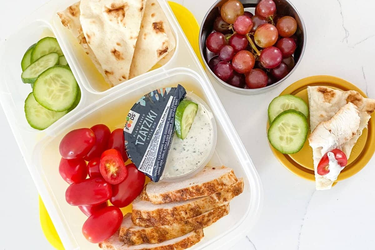 high protein greek chicken bento box with cherry tomatoes, sliced cucumber, pita and tzatziki sauce, plus grapes on a small container on the side.