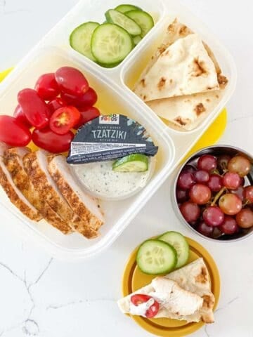 greek chicken bento box with cherry tomatoes, sliced cucumber, pita and tzatziki sauce, plus grapes on a small container on the side.