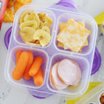 turkey, sliced cheese, baby carrots, and banana chips inside a snack box resembling a healthy lunchable