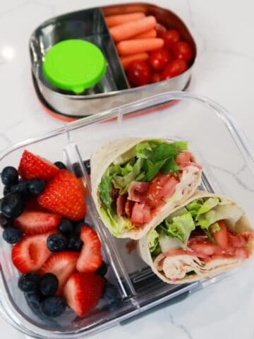 BLT wraps in a lunchbox with blueberries and strawberries on the side, and a smaller lunchbox with veggies behind