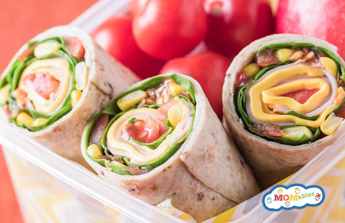 ham and veggie wraps in a lunch container with cherry tomatoes and an apple