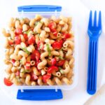 small square school lunch container filled with tomato pasta salad. Small blue plastic spoon next to the lunch.