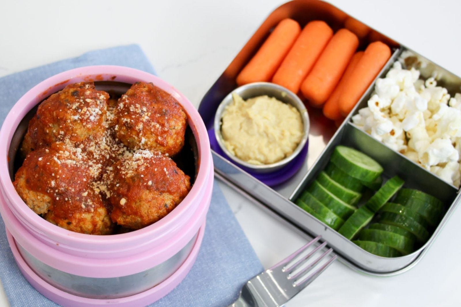 a thermos filled with hot meatballs in tomato sauce ready to be packed for school in a lunchbox next to a small snack box container with carrots, hummus, cucumbers, and popcorn for a complete lunch