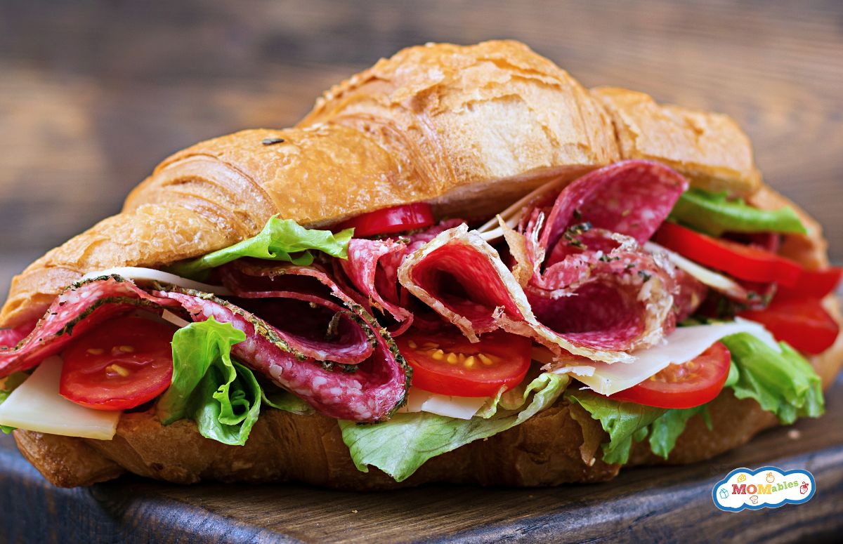 a croissant sandwich topped with lettuce, tomatoes, salami and cheese