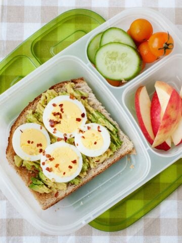 top view of a lunchbox with avocado toast, a sliced apple, nad veggies